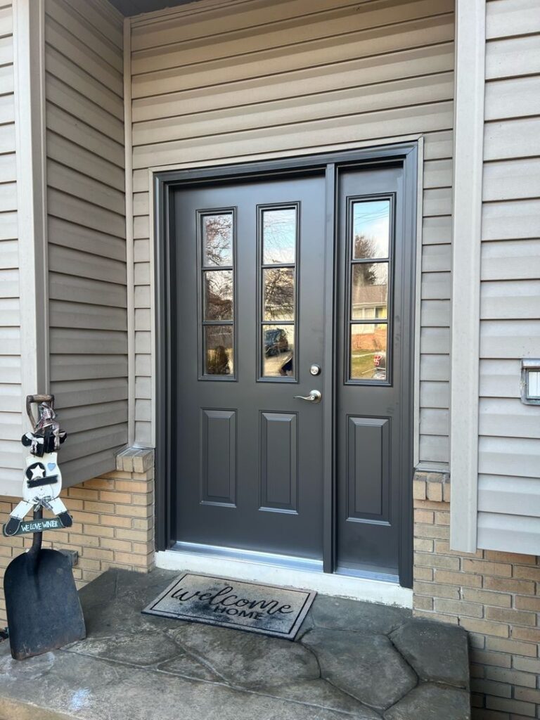 Gray front door with three vertical panels and glass side windows, surrounded by beige siding and adorned with a 'welcome home' mat