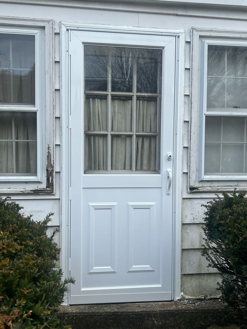 A new white storm door with upper window and lower panels on an older house with weathered trim and adjacent windows.